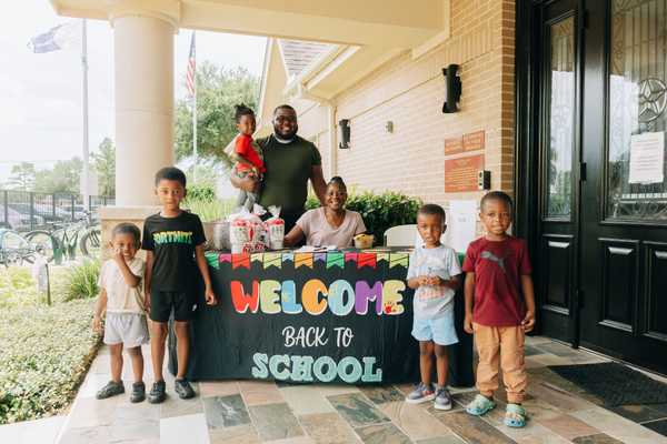 Group of children and adults gather around a welcome table with a “Back to School” sign outside a community building during a school readiness event.